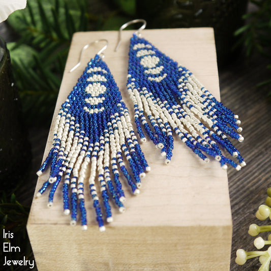 Blue and matte silver-white beaded moon phase fringe earrings on a wooden block with 'Iris Elm Jewelry' branding.