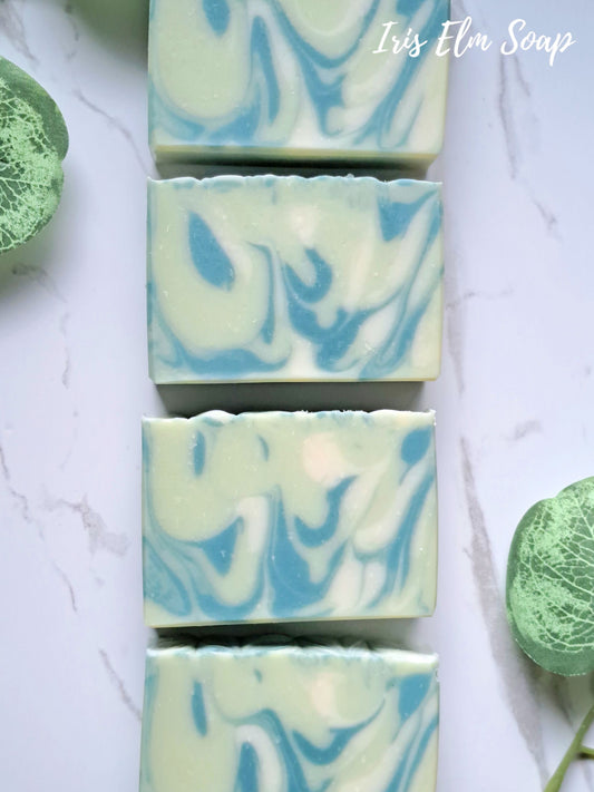 Four rectangular bars of swirled light green and blue agave lime handmade soap arranged vertically on a white marble surface, with decorative green leaves at the corners and the text 'Iris Elm Soap' in the top right.