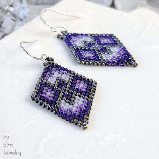 Pair of beaded purple diamond shape seed bead earrings with a geometric pattern on a white surface, branded 'Iris Elm Jewelry'.