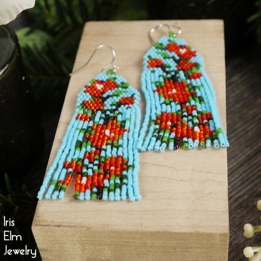 Colorful beaded light blue and red flower-patterned seed bead fringe earrings on a wooden block with 'Iris Elm Jewelry' branding.
