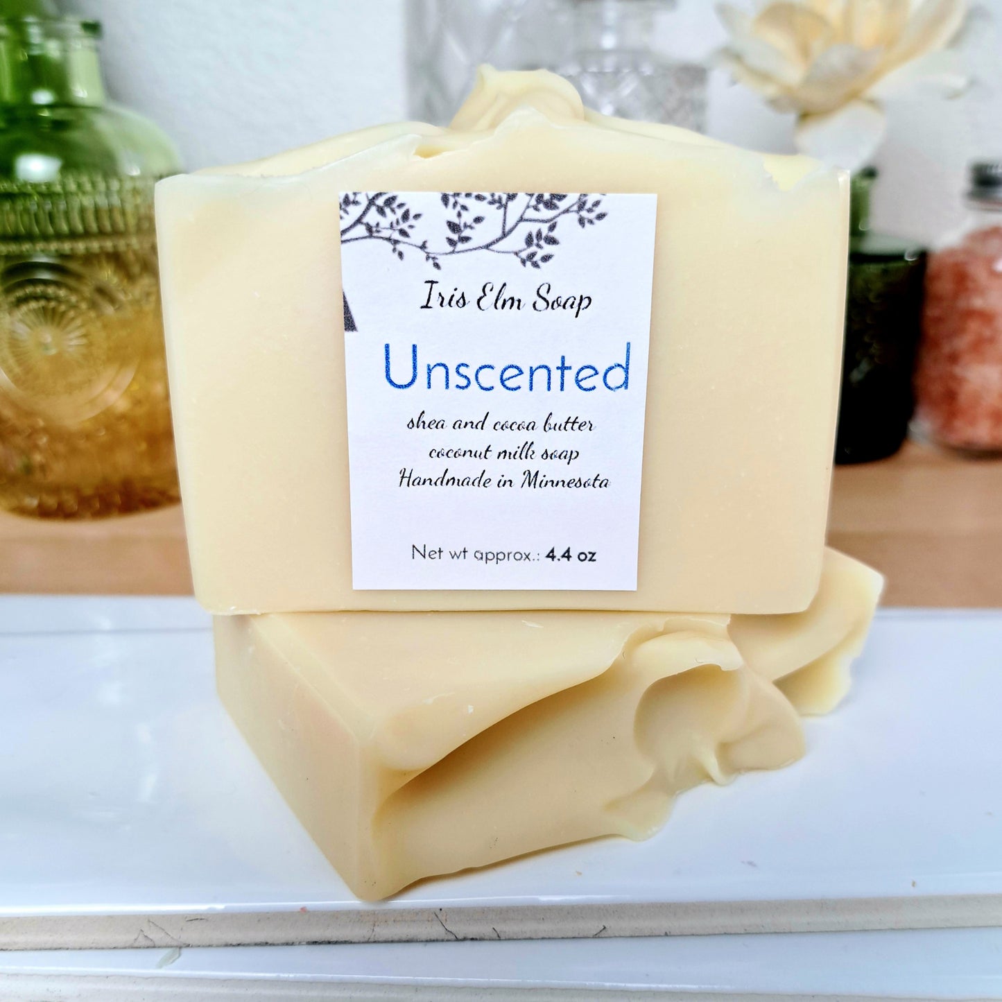 Unscented soap bar with a Iris Elm Soap label on a white surface, with a blurred background of bottles and flowers.