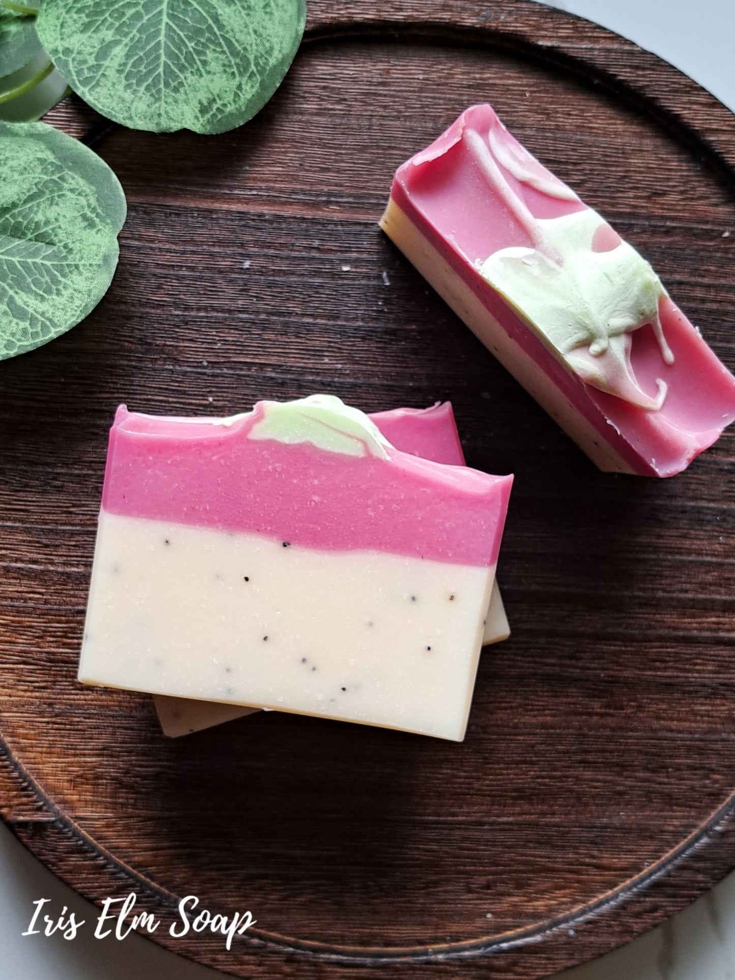 Two layered artisanal handmade pink dragonfruit soap bars on a dark wooden tray, with one bar showing a pink top layer over a creamy off-white bottom layer with tiny black specks, and the other showing a pink top with a pale green marbled swirl. The brand name 'Iris Elm Soap' is visible in the bottom left corner.