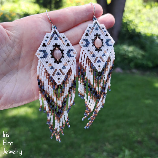 Beaded peach moonstone celestial moon cream, grey, copper seed bead diamond fringe earrings held in a hand with a blurred green outdoor background