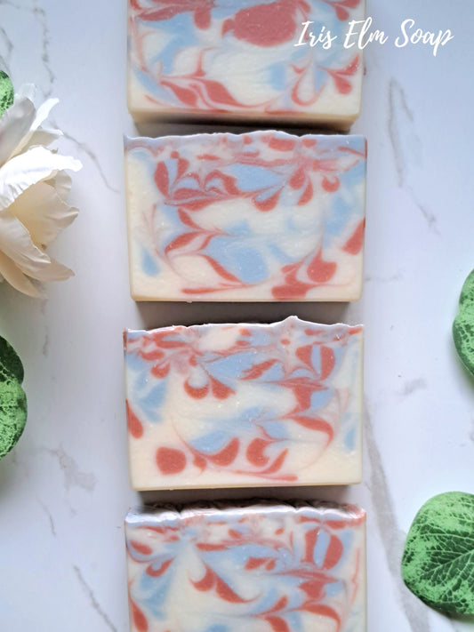 Four rectangular bars of swirled soap with cream, light blue, and coral-red marbling, arranged vertically on a white marble-like surface with decorative green leaves and a white flower. The text 'Iris Elm Soap' appears in the top right.