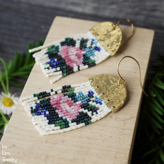 Beaded pink rose seed bead fringe earrings with hammered half circle brass and gold hooks on a wooden surface, surrounded by greenery.