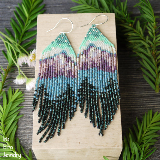 Beaded mountain lake forest seed bead fringe earrings on a wooden stand with greenery around