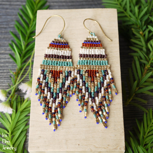 Beaded glass gold, blue, white, and purple seed bead striped fringe earrings on a wooden block with green leaves in the background