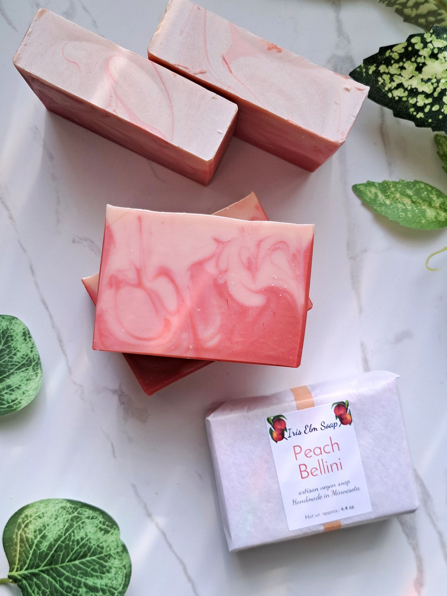 Pink soap bars with a 'Peach Bellini' label on a marble surface with green leaves.