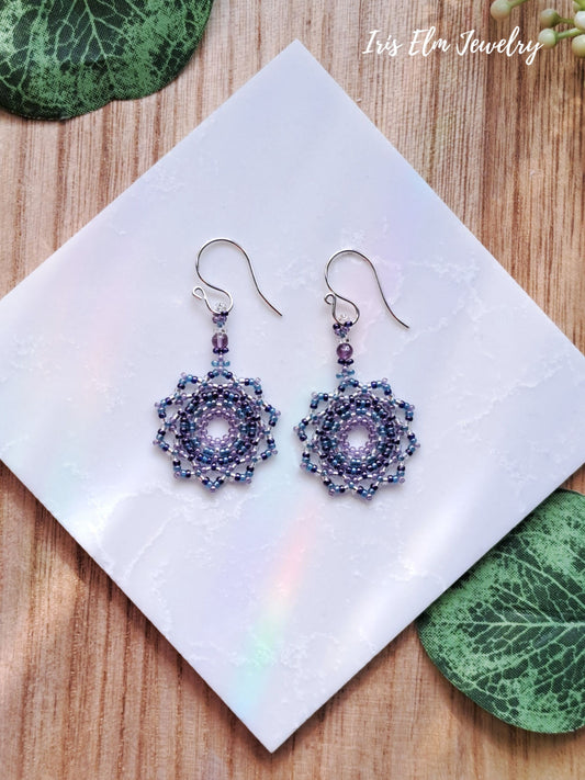 Purple beaded seed bead mandala earrings on a white square with green leaves around