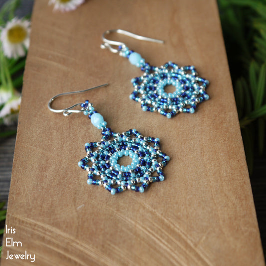 Blue beaded seed bead mandala earrings on a wooden surface with 'Iris Elm Jewelry' branding.