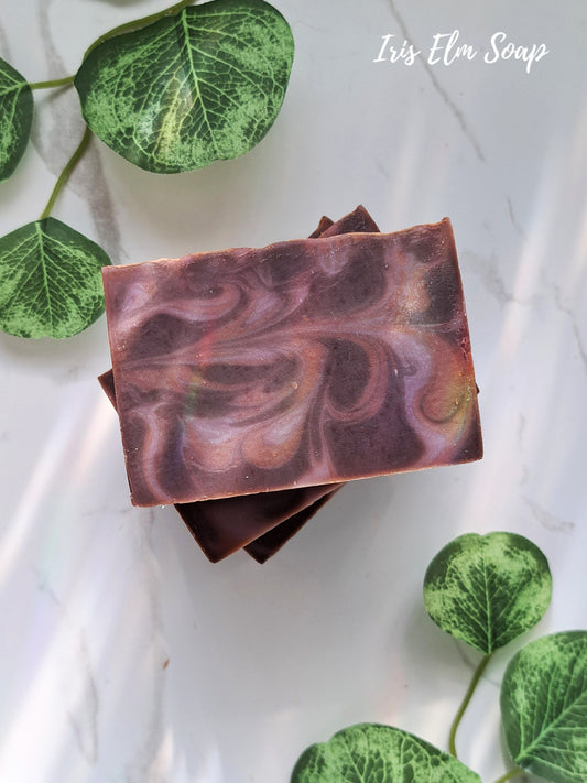 Two swirled purple and brown black amber and lavender handmade soap bars on a white background with green leaves.