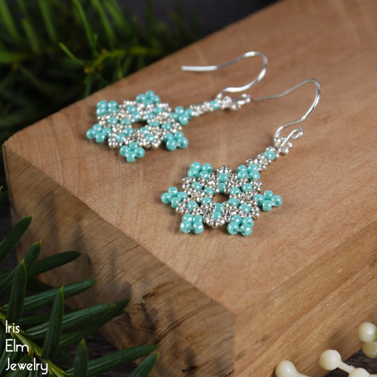 Turquoise glass seed bead beaded snowflake earrings on a wooden block with 'Iris Elm Jewelry' branding.