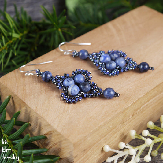 Blue beaded sodalite stone earrings on a wooden block with greenery in the background