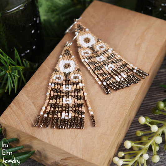 Beaded floral fringe seed bead earrings with neutral brown and white beads on a wooden block with greenery in the background