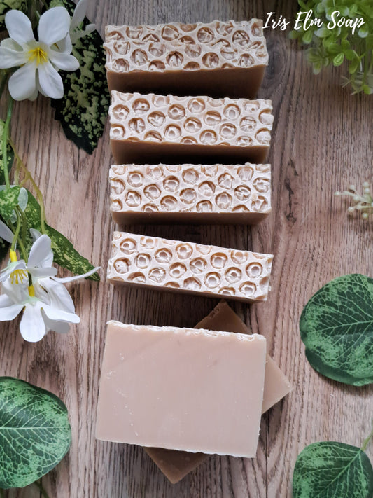 Beige oatmeal honey handmade soap bars with honeycomb pattern on a wooden surface with green leaves and flowers.