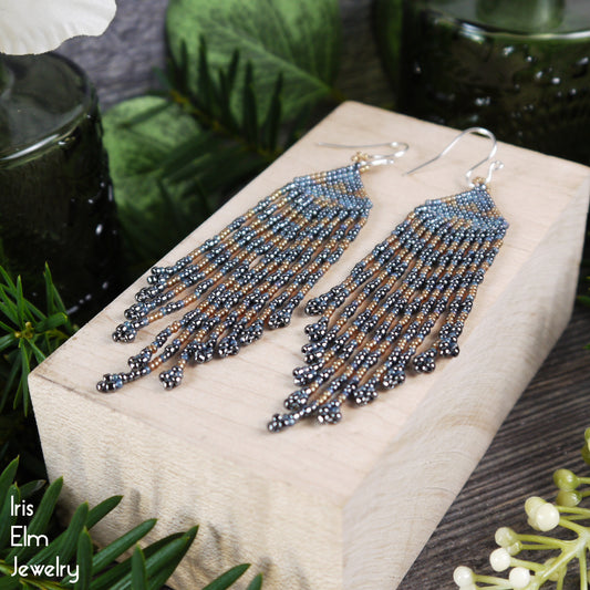 Beaded grey blue golden seed bead fringe earrings on a wooden block with plants in the background, featuring 'Iris Elm Jewelry' branding.
