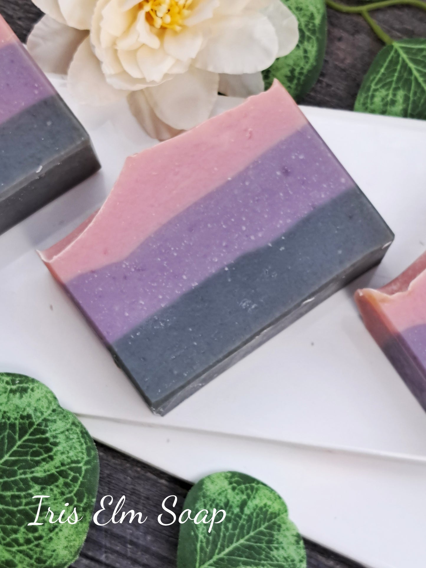 Three layered soap bars with 'Iris Elm Soap' branding on a white surface with green leaves.