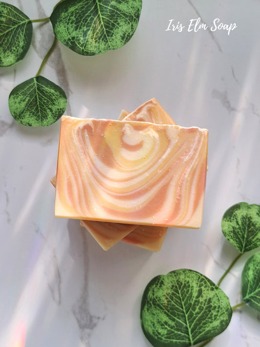 Two swirled vegan mango tangerine handmade soap bars on a marble surface with green leaves