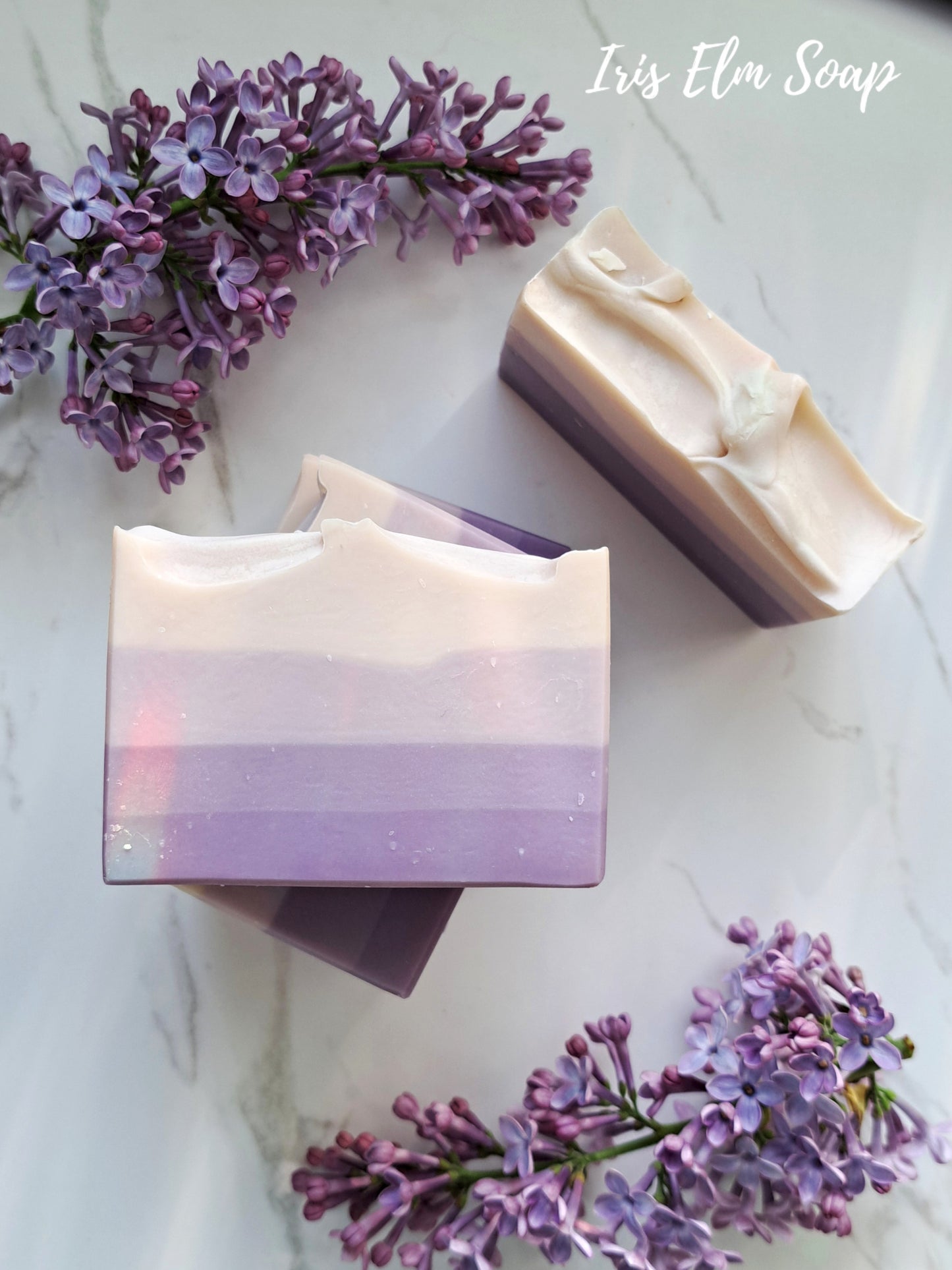 Two bars of French lilac handmade soap with a gradient design surrounded by purple flowers on a light background, with 'Iris Elm Soap' branding.