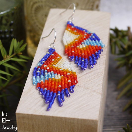 Colorful beaded red, orange, blue, yellow glass seed bead fringe earrings on a wooden block with 'Iris Elm Jewelry' branding.