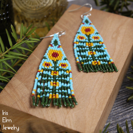 Beaded yellow sunflower seed bead fringe earrings on a wooden surface with greenery in the background, featuring the brand 'Iris Elm Jewelry'.