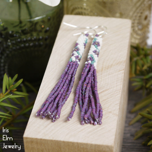 Pair of beaded purple and white flower tassel seed bead fringe earrings on a wooden block with 'Iris Elm Jewelry' branding.