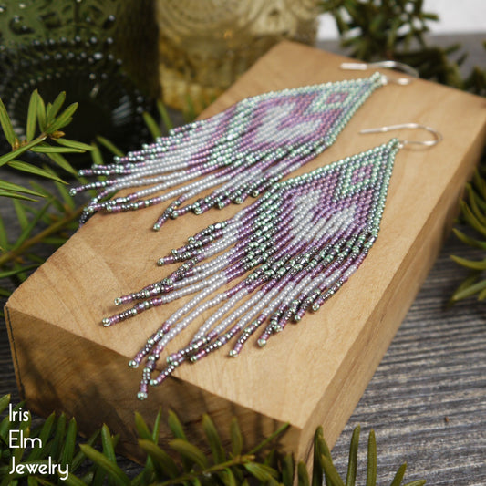 Beaded seafoam green, grey, and purple long seed bead fringe earrings on a wooden block with greenery in the background, featuring the brand 'Iris Elm Jewelry'.