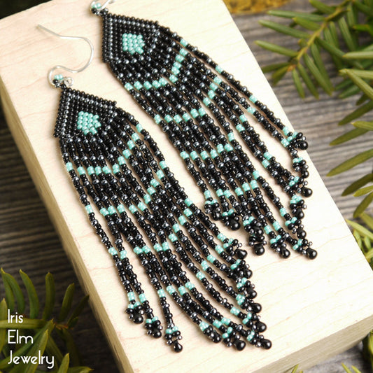 Black and turquoise beaded striped fringe seed bead earrings on a wooden block with greenery in the background