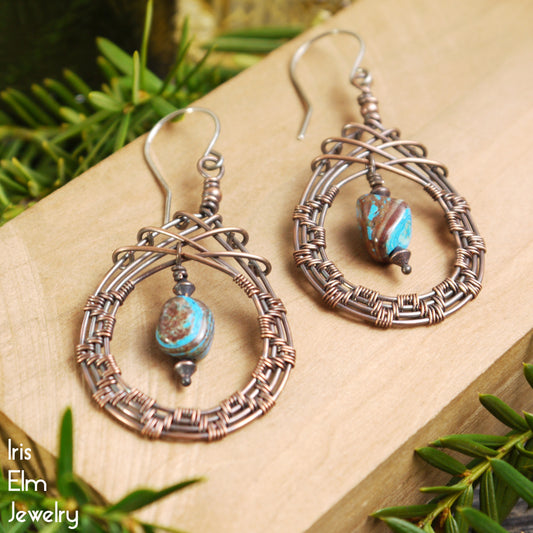 Pair of handmade blue sky jasper copper wire woven teardrop on a wooden surface with greenery.