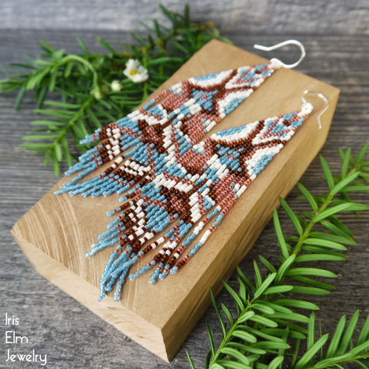 Beaded long fringe seed bead earrings with blue, white, and topaz brown geometric pattern on a wooden block with greenery around