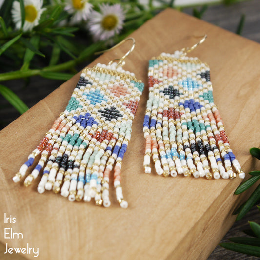 Colorful beaded seed bead diamond pattern fringe earrings on a wooden block with daisies in the background, featuring 'Iris Elm Jewelry' branding.