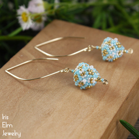 Pair of beaded triangle seed bead triangle blue white gold earrings with marquise goldfilled ear wires on a wooden surface with a blurred natural background