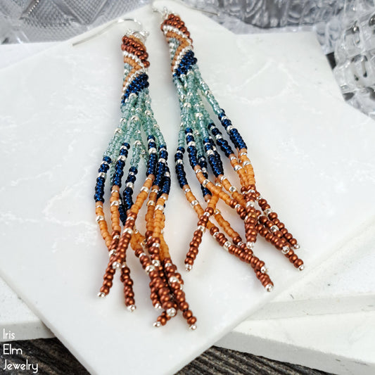 Beaded seed bead tassel fringe earrings with blue, orange, and silver beads on a white surface.