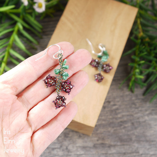 Hand holding a pair of beaded garnet and aventurine handmade flower earrings with a wooden block and greenery in the background