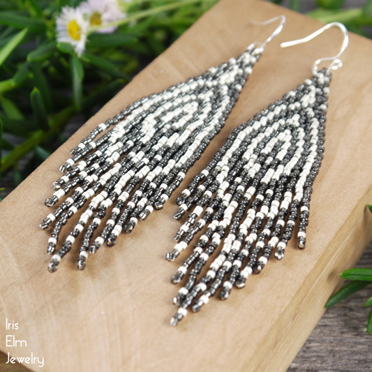 handmade beaded grey and matte silver white seed bead zebra pattern fringe earrings on a wooden surface with a natural background