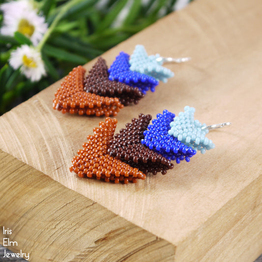 Colorful beaded blue and rust brown four tier triangle seed bead earrings on a wooden surface with blurred flowers in the background