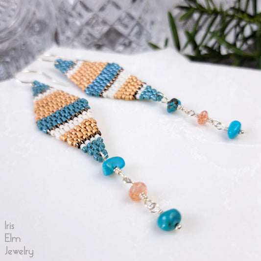 kite shaped beadwork seed bead earrings with blue and orange stripes with turquoise gemstone and sunstone dangling beneath on a white surface