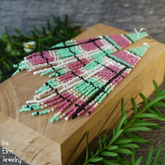 Colorful beaded seed bead plaid patterned fringe earrings on a wooden block with greenery in the background, featuring 'Iris Elm Jewelry' branding.