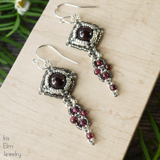 Pair of silver and dark red garnet beaded art nouveau earrings on a wooden surface with 'Iris Elm Jewelry' branding.