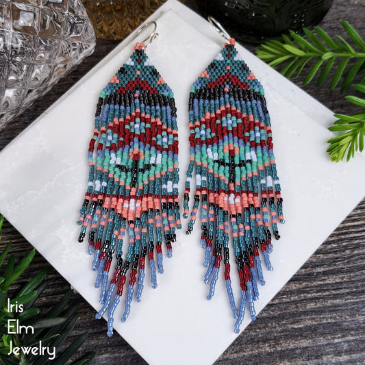 Beaded red, blue, mint, and coral floral folk art seed bead fringe earrings on a white square with 'Iris Elm Jewelry' branding, surrounded by greenery.
