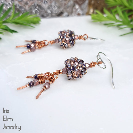 Pair of beaded purple glass and copper earrings on a white surface with greenery in the background, featuring 'Iris Elm Jewelry'.