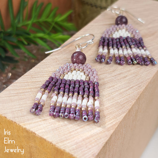 Beaded lepidolite lilac purple small seed bead and stone fringe earrings on a wooden block with a plant in the background, featuring the brand 'Iris Elm Jewelry'.