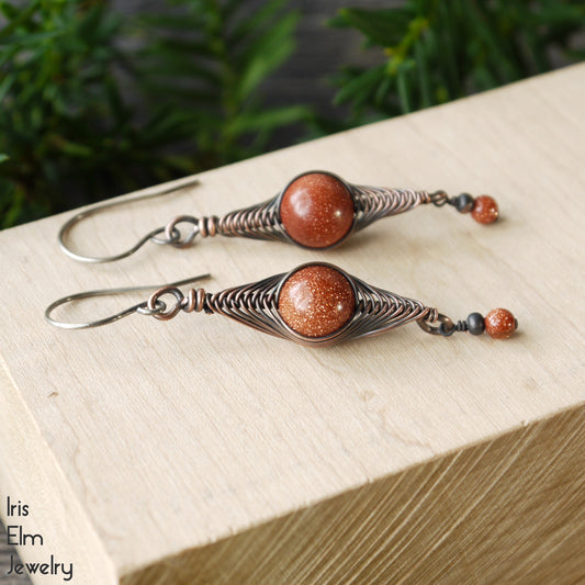 Pair of copper wire wrapped herringbone earrings with brown goldstone beads on a wooden block, brand 'Iris Elm Jewelry' visible.