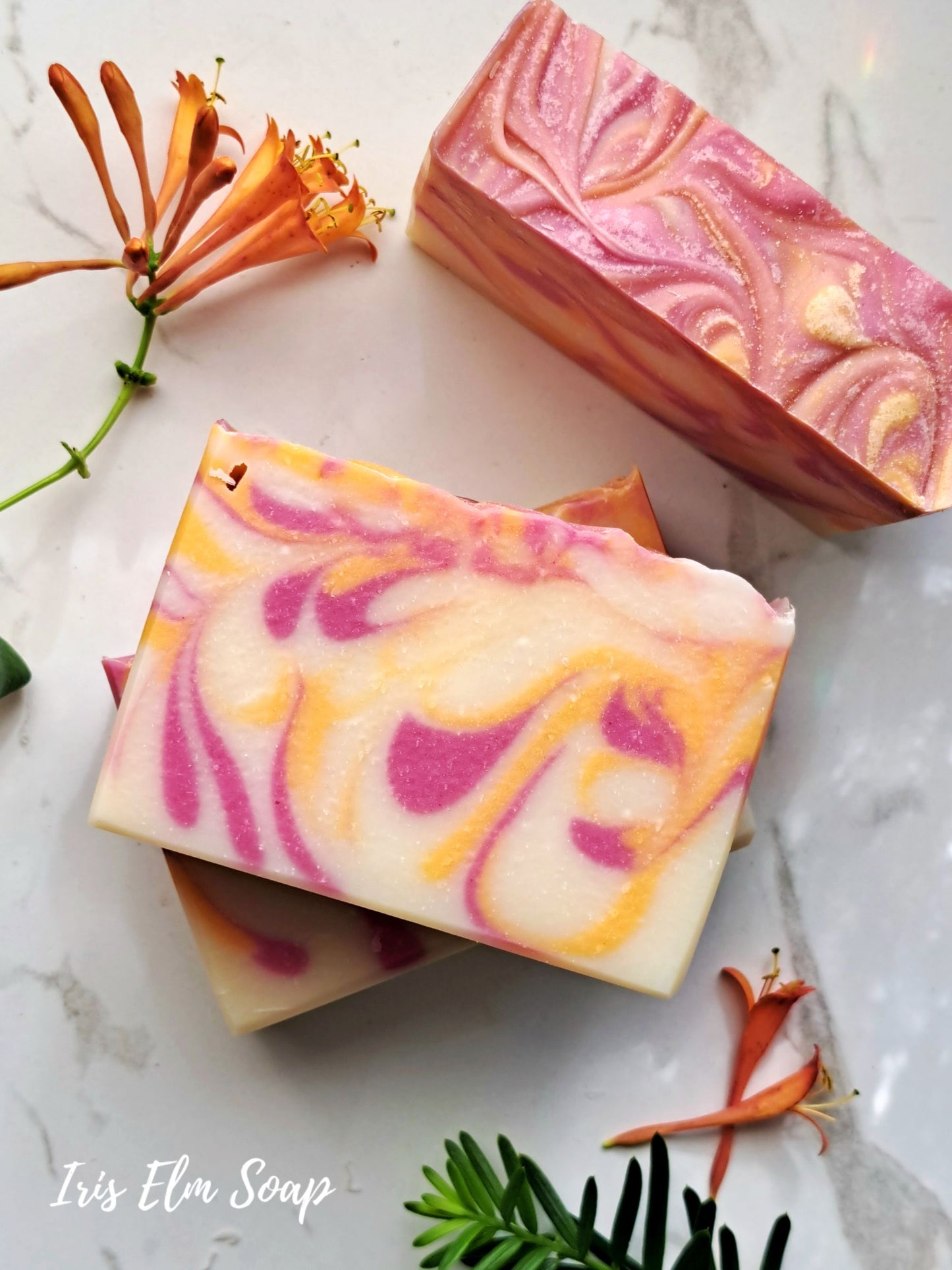 Swirled honeysuckle handmade soap bars with pink and orange hues on a marble surface with flowers and greenery.