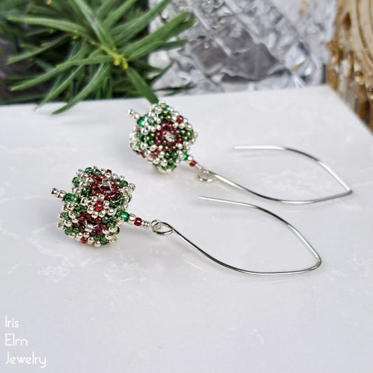 Pair of beaded cube glass seed bead marquise drop earrings with green and red beads on a white surface with a blurred natural background.