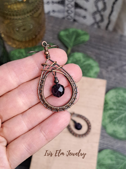 Copper wire-wrapped earring with a dark purple amethyst stone bead held in hand, on a textured surface.