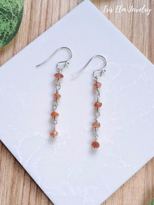 A pair of dangle earrings with silver-toned fishhook wires and small peach-orange faceted sunstone gemstone beads separated by tiny silver links, displayed on a white square card over a wood surface. The text 'Iris Elm Jewelry' appears in the top right.