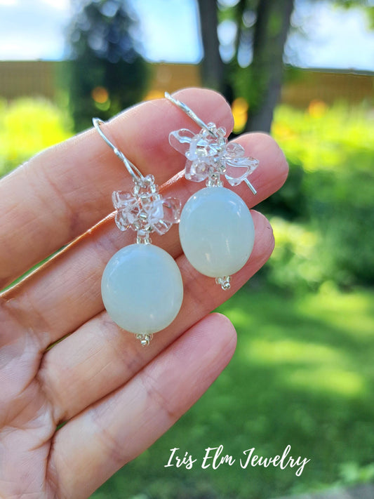 A hand holding a pair of dangling earrings outdoors with a blurred green garden background. The earrings have silver-toned metal hooks and floral wire details with oval milky seafoam green serpentine jade beads hanging below.