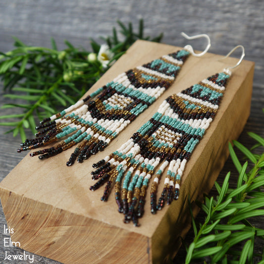 Long beaded seed bead brown and blue fringe earrings on a wooden block with greenery in the background, featuring the brand 'Iris Elm Jewelry'.