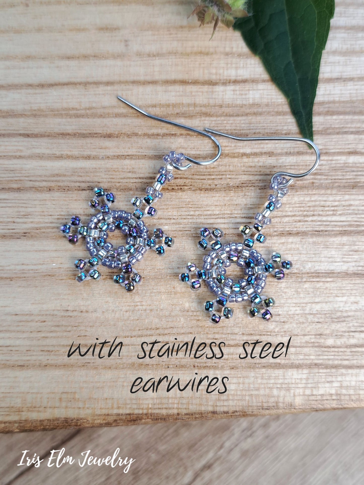 A pair of handmade beaded dangle earrings with silver-toned stainless steel ear wires, displayed on a light wood background with a green leaf. The text overlay reads 'with stainless steel earwires' and the brand 'Iv's Elm Jewelry' is visible in the bottom left corner.
